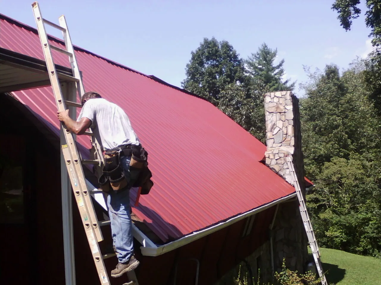 Roofer on a ladder beside a red metal roof and stone chimney — Soffit & Fascia Repair by Roofing Brothers in Hazen